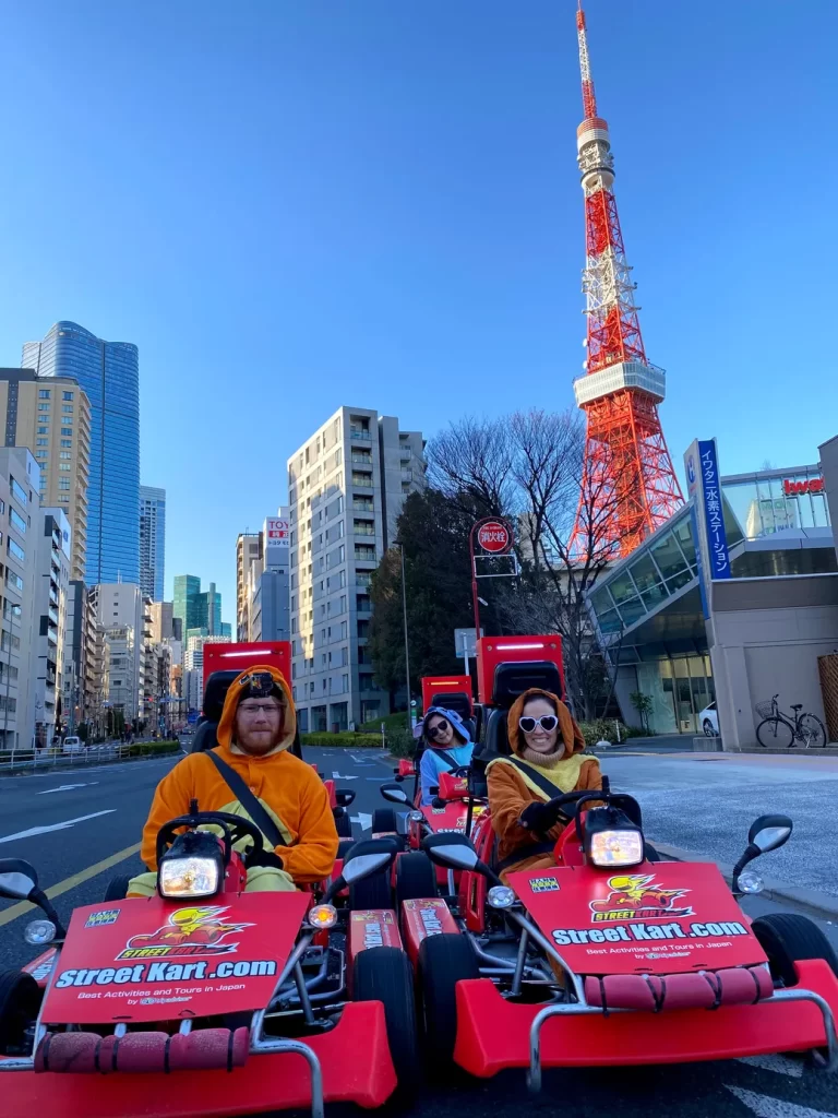IMG_3639 - StreetKart People in orange costumes drive red go-karts on a city street, Tokyo Tower visible in the skyline behind them.