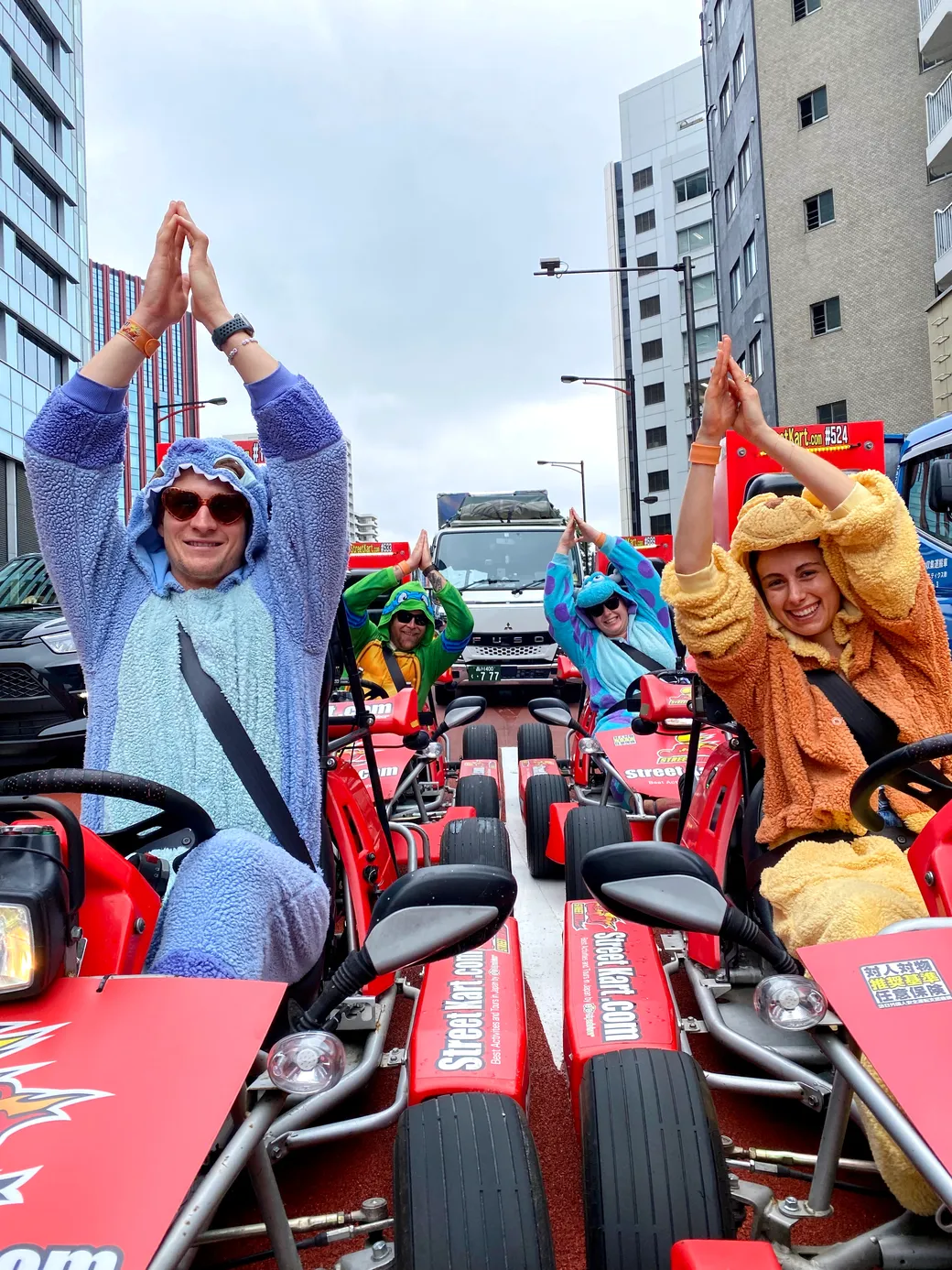Group of people in colorful animal costumes posing in go-karts on a city street, hands together in a prayer-like position as they smile at the camera